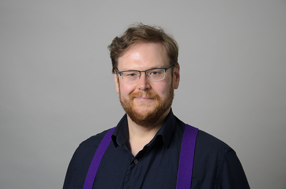 Portrait of Luke Reynolds wearing a black collared shirt with purple suspenders.